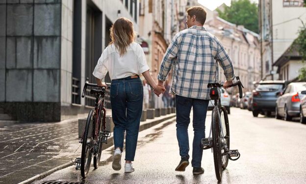 Photo of back view of couple holding hands and walking with bicycles on city street after rain. Concept of addiction and relationships